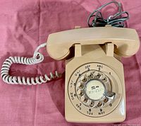 Beige Western Electric rotary dial phone lying on a pink fabric surface, showing handset and coiled cord connected to base with rotary dial visible.