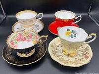Four English fine bone china teacups with matching saucers arranged on a black background, showing distinct colors and gold gilt detailing.