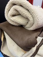 Close-up view of beige faux shearling fabric rolled, showing texture of soft curly pile; brown faux leather-backed faux shearling fabric underneath.