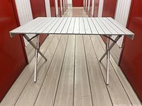Full view of lightweight aluminum roll-top folding table set up in hallway, showing the parallel slats of the table top and legs extended.