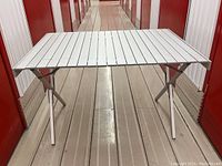 Full view of the aluminum roll-top table set up, showing the slatted top and foldable legs, photographed in a narrow corridor.