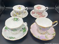 Four Royal Albert bone china teacups and matching saucers showing floral patterns and gold gilt accents, set arranged on black background.