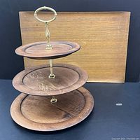 3-tier wooden serving tray with brass hardware and rectangular teak veneer tray behind it on black background.