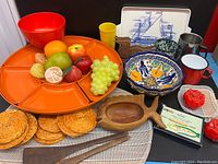 Overall lot showing colorful vintage serving ware including orange segmented lazy Susan, red mixing bowl, Mexican ceramic bowl, blue and white tray, enamelware cups, kitschy fruit candles, artificial fruit, woven orange coasters, wooden decor and utensils.