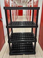 Full view of assembled black plastic storage shelf showing four grid-pattern shelves in a narrow hallway.