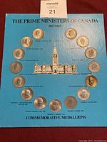 Blue display board titled 'THE PRIME MINISTERS OF CANADA 1867-1969' with 14 circular medallions arranged in a semi-circle above an illustration of the Canadian Parliament building. Each medallion displays a relief portrait of a different Prime Minister and is labeled with their name and years of service.