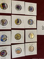 Twelve USA State Quarter Dollar coins with colored designs, each in protective holders, laid out on a red surface.