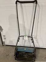 Front view of manual push lawn mower showing gray metal frame, black handlebars, and blue blade cover.