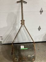 Full view of a large metal lawn roller with a cylindrical drum and long handle against a white garage door. The metal surface shows rust and wear.