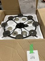 Photo showing the metal gazebo chandelier inside an open cardboard box with protective foam packaging, instruction manual visible in the background, chandelier placed upside down showing bottom side with candle holders and hanging chains.