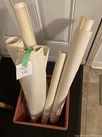 Photo shows rolled maps placed vertically in a red crate against a door, multiple maps of varying diameter visible, some secured with green tape, showing aged, off-white paper typical of older topographic maps.