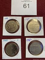 Photo of four one dollar coins from Canada and the US in individual holders with handwritten dates and notes on a red background.