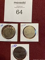 Photo of three Canadian one dollar coins from years 1969, 1979, and 1982 placed on a red surface with auction lot number 64 card