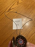 Open pocket watch showing a black face with white numbers and a Captain America emblem in the center, along with attached silver chain.