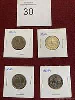 Four USA quarter dollar coins in individual white cardboard holders, each marked with 'USA' in blue ink. Coins show the eagle on the reverse side. One coin shows notable wear and discoloration.