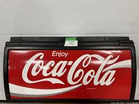 Rectangular red and white Coca-Cola sign framed in black plastic, featuring the classic Coca-Cola script logo in white.