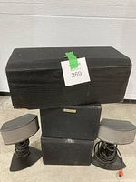 Five black speakers stacked in two columns: two small curved bookshelf speakers with cables on a stand at top left and right, one mid-sized rectangular speaker with fabric cover below them, and two larger rectangular speakers with fabric covers stacked in the middle and bottom right.