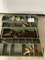 Open green metal fishing tackle box showing three layers of compartments filled with various fishing lures and hooks.