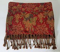 Close-up of folded floral patterned table runner showing red base with gold and green flowers and brown accents, including fringed twisted tassels at the edge.