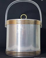 Front view of the ice bucket showing brushed silver tone body, gold accent bands on top and bottom edges, and clear Lucite handle with gold loop detail on lid.