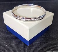 Silver-tone plated stainless steel bangle bracelet with a row of small clear Austrian crystals, displayed on a white and blue box against a black background, showcasing the bracelet's front view and sparkle.