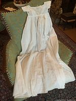 Full length view of the antique white christening gown laid on a green upholstered chair showing the full length, fabric drape, and lace details.