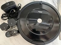 Top-down view of the black iRobot Roomba vacuum cleaner with four accessory items: charging dock with cable, remote control, and two power adapters.