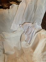 Six white and off-white antique and vintage baby clothes laid flat showing pintucks, embroidery, lace trims and smocking detail.