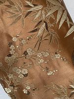 Close up of bronze silk satin fabric showing detailed floral and bamboo brocade patterns in gold and cream tones.