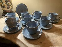 Photo showing eight periwinkle blue ceramic cups paired with matching saucers arranged on a surface.