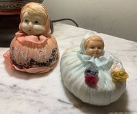 Photo displaying two vintage pin cushions with celluloid doll heads dressed in fabric bodices, one in peach embroidery and the other in light blue with fabric flowers.