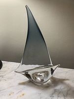 Side view of art glass sailboat figurine with tall gray sail and clear glass body on marble surface.