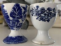 Two blue and white egg cups, one with a detailed leaf and landscape design, the other with a floral motif, shown side by side.