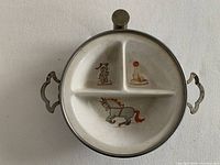 Top view of the vintage metal baby warming dish showing three compartments with circus themed illustrations including a clown, an elephant, and a horse. Dish has two side handles.