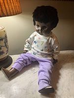 Full view of vintage black doll sitting on marble surface next to a lamp, showing clothing and doll features including hair and feet.