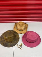 Three hats displayed side by side showing variety in styles and colors: pink wicker hat, natural straw wicker hat with turquoise beads, brown cowboy style hat with embossed designs.