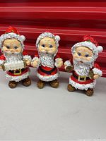 Front view of three papier-mâché Santa figures each about 8.5 inches tall with textured white trim, red cloaks, and gold accents.