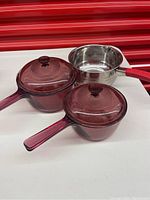 Top view showing two cranberry Pyrex pots with lids and one stainless steel pot with red handle