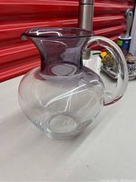 Front angled view showing clear round body with violet-tinted neck and top rim, and clear handle attached on side.