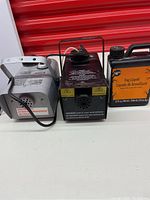 Two fog machines side by side with fog liquid bottle on white surface.