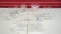Wide view showing a varied collection of different types of glassware and crystal including wine glasses, tumblers, stemmed glasses, and cups arranged on white surface.