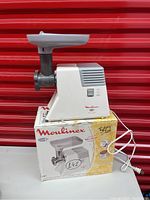 Electric grinder unit placed on original Moulinex box, showing on/off switch, power cord, and vented top with product branding.