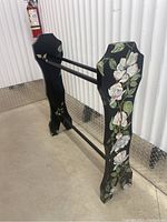 Angled side view of the handpainted black wood quilt rack showing floral decoration on the side panel and two horizontal rods.