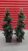 Front view of two decorated artificial Christmas trees in dark brown square bases with lights, pinecones, and red berries, standing side by side.