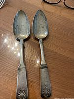 Pair of two antique silver serving spoons photographed on a wooden surface with eyeglasses in background.