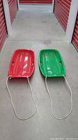 Overview of two Slippery Racer sleds, one red and one green, lying on concrete. Both have attached white pull ropes.