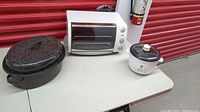 Oster toaster convection oven, Proctor-Silex rice cooker, and black-speckled roaster pan on a folding table