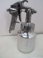 Side view of metal paint sprayer with attached paint container and trigger, showing overall finish and condition.