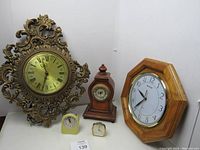 Photo showing all six clocks arranged together: large ornate metal framed wall clock, octagonal oak wall clock, wooden case table clock, two small alarm clocks, yellow Westclox travel clock missing case.