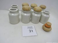Eight white ceramic containers, six with wooden stoppers, two without, arranged in two rows with one stopper loose on the surface.